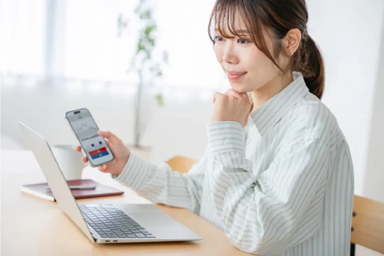 女性がパソコンの前に座りスマホ片手に考えている写真